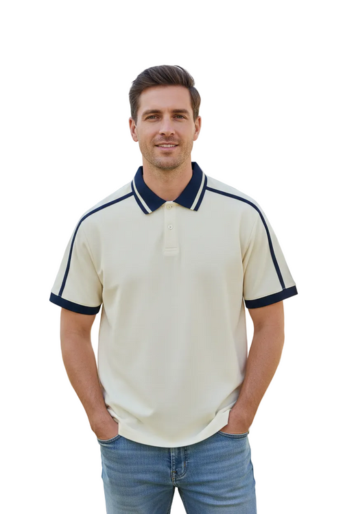 A man wearing a cream-colored short-sleeve polo shirt with navy blue trim on the collar and sleeve cuffs, paired with blue jeans, standing outdoors in front of a modern building and greenery.